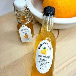 May include: Two glass bottles of pure orange extract with a white label that says "Made with Love Pure Orange Extract". The label also includes a small orange illustration. The larger bottle is filled with a light orange liquid and has a black cap. The smaller bottle has a white cap and a clear glass stopper.