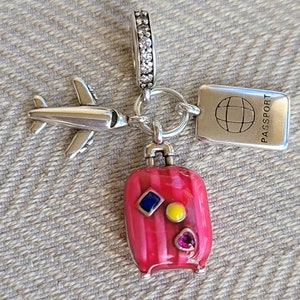 May include: Silver charm with a pink suitcase, a silver airplane, and a silver tag that says "PASSPORT" with a globe symbol.