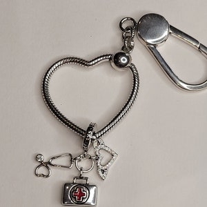 Puede incluir: Llavero de plata en forma de corazón con un dije de plata de una bolsa médica con una cruz roja, un estetoscopio y un dije de corazón.
