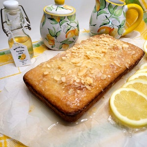 May include: A loaf of lemon cake topped with sliced almonds and a lemon glaze. The cake is sitting on a white parchment paper on a yellow and green checkered tablecloth. There is a small bottle of pure lemon extract, a lemon-themed sugar bowl, and a lemon-themed creamer next to the cake.
