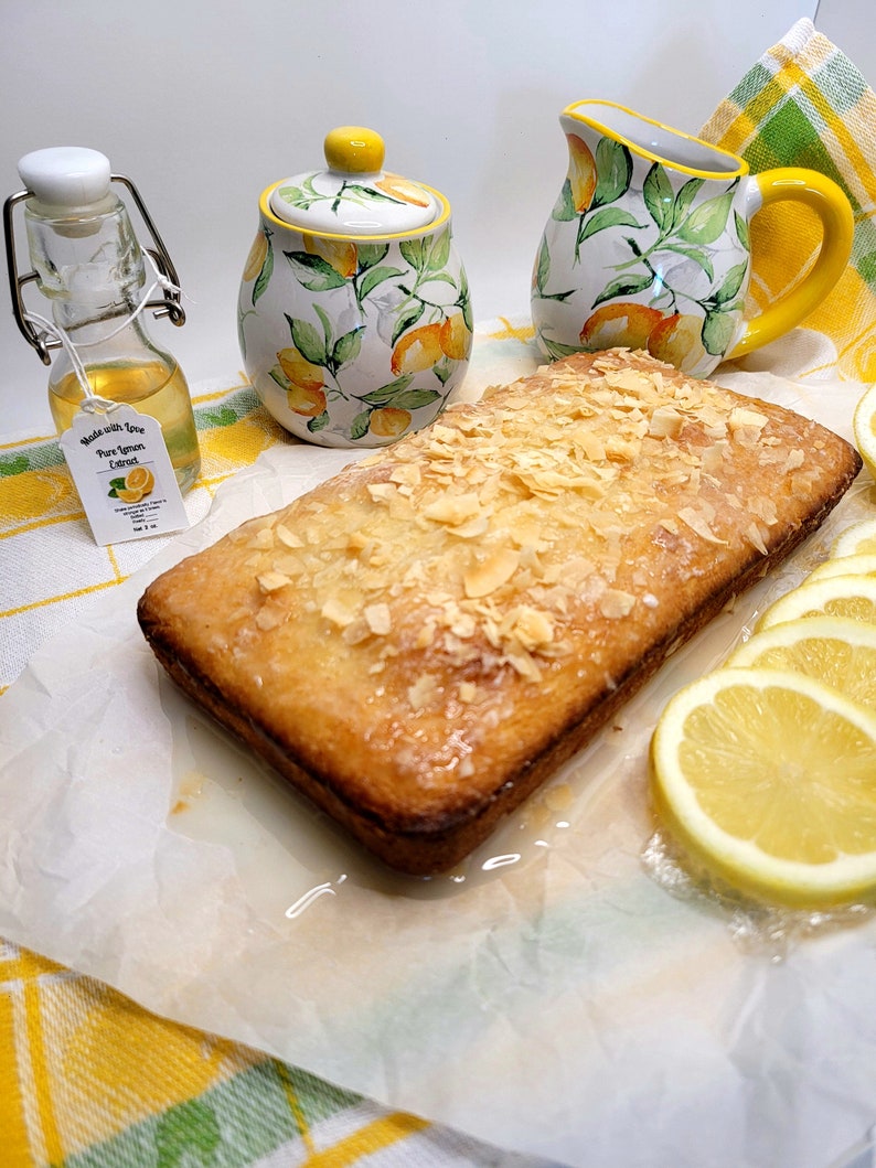 May include: A loaf of lemon cake topped with sliced almonds and a lemon glaze. The cake is sitting on a white parchment paper on a yellow and green checkered tablecloth. There is a small bottle of pure lemon extract, a lemon-themed sugar bowl, and a lemon-themed creamer next to the cake.