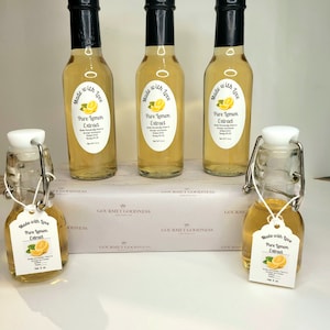 May include: Five glass bottles of pure lemon extract, labeled "Made with Love, Pure Lemon Extract". The bottles are arranged in a row on a white surface with a white box in the background. The box has the text "Gourmet Goodness" printed on it.