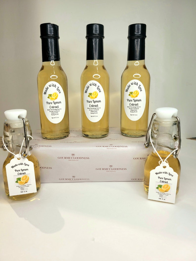 May include: Five glass bottles of pure lemon extract, labeled "Made with Love, Pure Lemon Extract". The bottles are arranged in a row on a white surface with a white box in the background. The box has the text "Gourmet Goodness" printed on it.