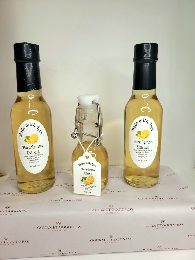 May include: Three glass bottles of pure lemon extract, two are larger and one is smaller. The bottles are clear and filled with a light yellow liquid. Each bottle has a black cap and a white label with the text "Made with Love Pure Lemon Extract" and a small lemon illustration.