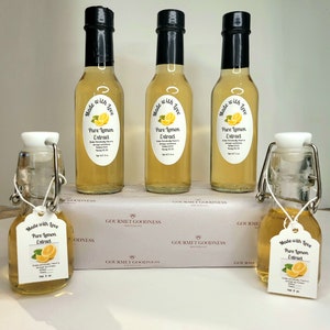 May include: Five glass bottles of pure lemon extract, each with a label that reads "Made with Love, Pure Lemon Extract". The bottles are arranged in a row on a white surface with a white box in the background. The box has the words "Gourmet Goodness" printed on it.