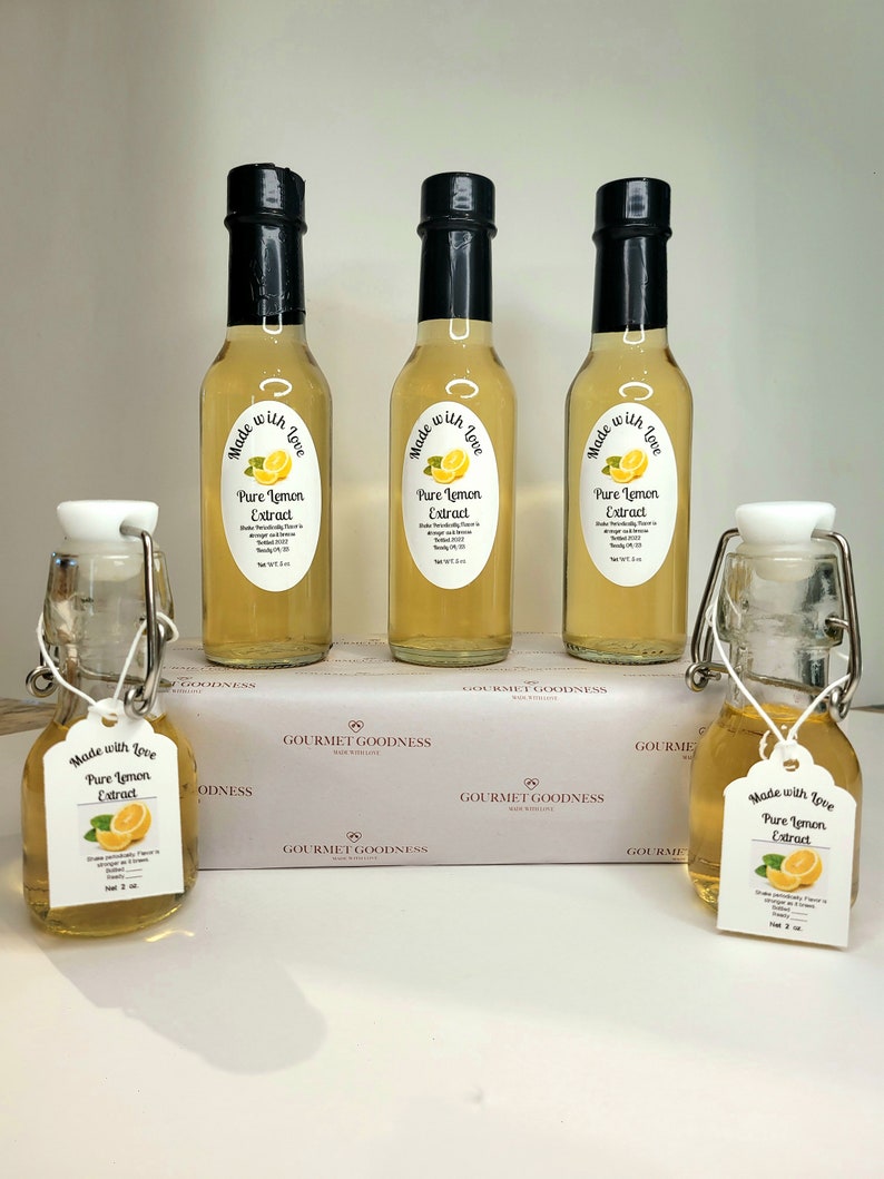 May include: Five glass bottles of pure lemon extract, each with a label that reads "Made with Love, Pure Lemon Extract". The bottles are arranged in a row on a white surface with a white box in the background. The box has the words "Gourmet Goodness" printed on it.