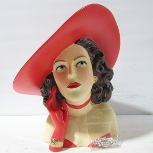 May include: A red ceramic bust of a woman wearing a large red hat and a red glove. The woman is wearing a red and white dress and a gold bracelet. The bust is a vintage style and is a decorative item.