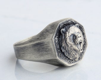 Könnte beinhalten: Ein silberner Ring mit einem detaillierten Bärenkopfdesign. Der Ring hat eine strukturierte, achteckige Band.