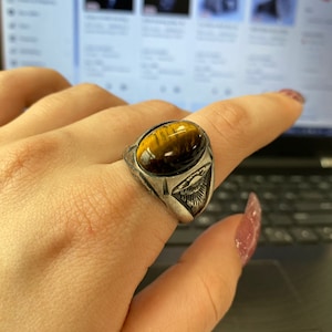 Op de afbeelding: Een zilverkleurige ring met een ovale tijgeroogsteen. De ring heeft een gedetailleerd ontwerp aan de zijkanten, met een adelaar- of vogelachtig motief. De steen heeft bruine en gouden kleuren.