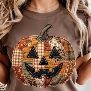 May include: A brown short-sleeved t-shirt featuring a large, central pumpkin graphic. The pumpkin is constructed from various fabric patches in orange, brown, and tan tones, including plaid, floral, and leopard prints, with black felt facial features.