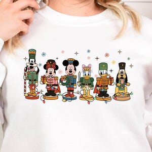 May include: White long-sleeve sweatshirt featuring a colorful graphic of Disney characters dressed as nutcrackers. The characters are arranged in a row, with festive lights and snowflake accents. A red Christmas ornament is held in the hand.