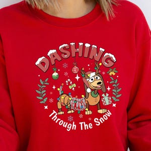 Może przedstawiać: Czerwona bluza z nadrukiem świątecznym. Projekt przedstawia kresk&oacute;wkowego psa z rogami i światełkami oraz napis "Dashing Through The Snow". Odzież świąteczna.