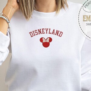 May include: White sweatshirt with the text "DISNEYLAND" in red and a red Mickey Mouse head with a bow.