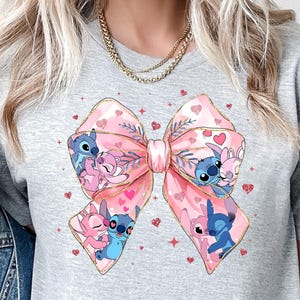 Puede incluir: Una camiseta gris claro con un gran dise&ntilde;o de lazo rosa con personajes de dibujos animados. El lazo est&aacute; decorado con corazones y los personajes son azules y rosas. La camiseta tiene mangas cortas.