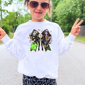 May include: A child wearing a white long-sleeved sweatshirt with a colorful graphic featuring characters. The child is also wearing black biker shorts with a white smiley face pattern and white socks. The child is wearing sunglasses and making a peace sign.