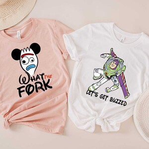 May include: Two white t-shirts with a pink and white background. The pink t-shirt has a cartoon image of a fork with Mickey Mouse ears and the text "What the Fork". The white t-shirt has a cartoon image of Buzz Lightyear with the text "Let's get buzzed".