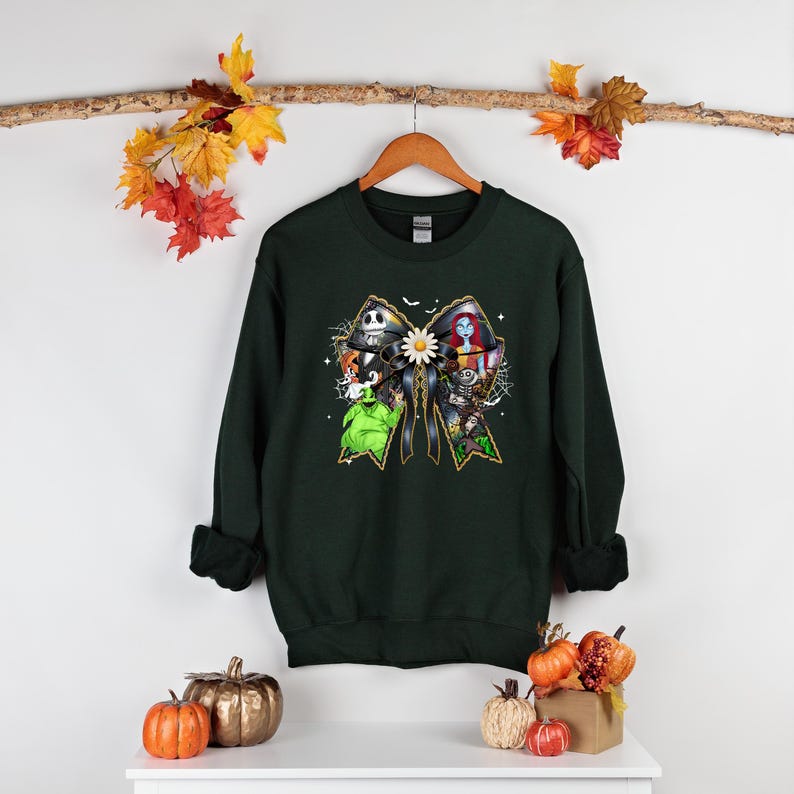 May include: Dark green sweatshirt featuring a colorful graphic of characters from The Nightmare Before Christmas arranged in a bow shape. The design includes Jack Skellington, Sally, and Zero. The sweatshirt is hanging on a wooden hanger.
