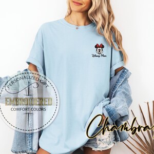 May include: Light blue t-shirt with a small embroidered Minnie Mouse design and the text "Disney More." The shirt has rolled-up sleeves and is part of the Comfort Colors collection. A denim jacket is worn over the shirt.