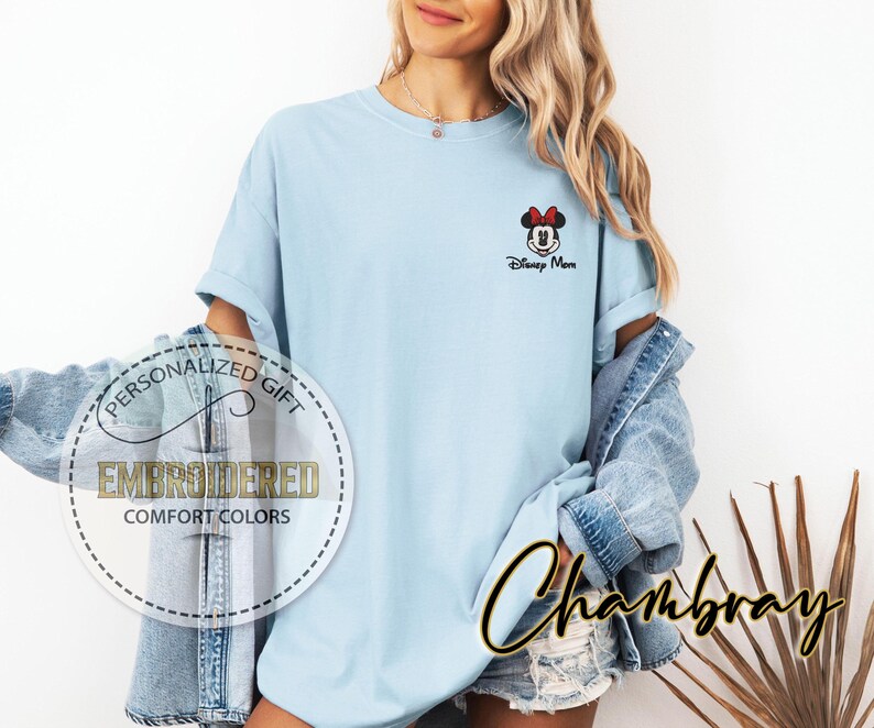 May include: Light blue t-shirt with a small embroidered Minnie Mouse design and the text "Disney More." The shirt has rolled-up sleeves and is part of the Comfort Colors collection. A denim jacket is worn over the shirt.