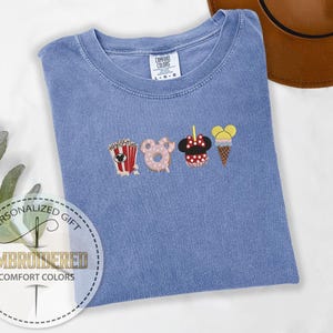 May include: A light blue t-shirt with embroidered images of popcorn, a donut, Minnie Mouse, and an ice cream cone. The text "COMFORT COLORS" is visible on the shirt.