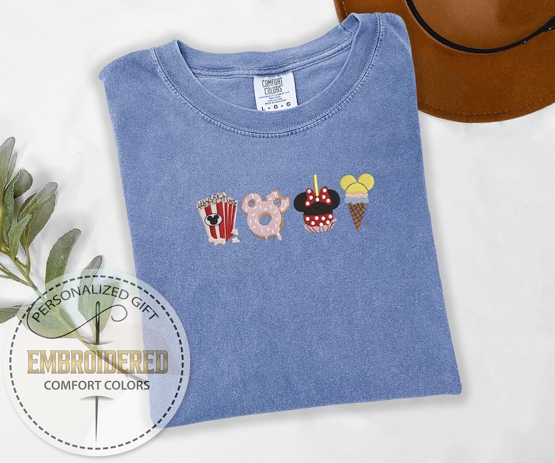 May include: A light blue t-shirt with embroidered images of popcorn, a donut, Minnie Mouse, and an ice cream cone. The text "COMFORT COLORS" is visible on the shirt.