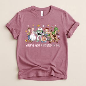 May include: Mauve t-shirt featuring a colorful graphic of Toy Story characters, including Woody, Buzz Lightyear, and others. The text "YOU'VE GOT A FRIEND IN ME" is printed below the characters. The shirt has rolled-up sleeves.