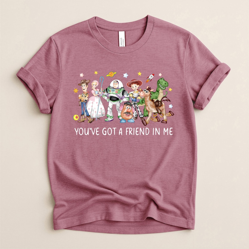 May include: Mauve t-shirt featuring a colorful graphic of Toy Story characters, including Woody, Buzz Lightyear, and others. The text "YOU'VE GOT A FRIEND IN ME" is printed below the characters. The shirt has rolled-up sleeves.