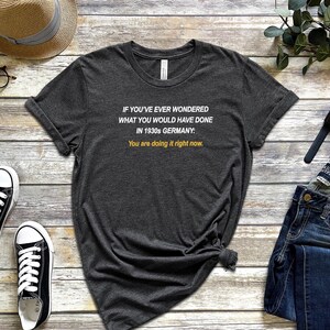 May include: A dark grey t-shirt with white text that reads "IF YOU'VE EVER WONDERED WHAT YOU WOULD HAVE DONE IN 1930s GERMANY: You are doing it right now."