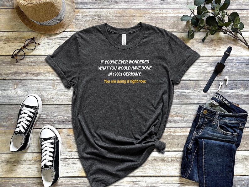 May include: A dark grey t-shirt with white text that reads "IF YOU'VE EVER WONDERED WHAT YOU WOULD HAVE DONE IN 1930s GERMANY: You are doing it right now."