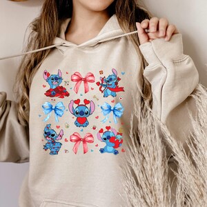 K&ouml;nnte beinhalten: Ein beiger Hoodie mit einem Grafikdruck eines blauen Comic-Charakters mit roten Herzen und Schleifen. Der Charakter ist in verschiedenen Posen dargestellt, darunter das Halten eines Herzens, das Schie&szlig;en eines Pfeils und das Tragen einer Schleife.