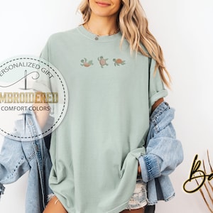 May include: A light green, oversized t-shirt with embroidered turtles. The shirt features the text "EMBROIDERED COMFORT COLORS" in a circular design. A denim jacket is draped over the shoulders.