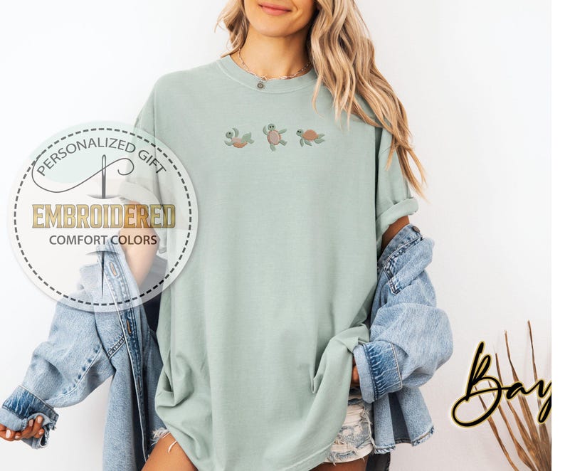 May include: A light green, oversized t-shirt with embroidered turtles. The shirt features the text "EMBROIDERED COMFORT COLORS" in a circular design. A denim jacket is draped over the shoulders.