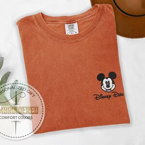 May include: Burnt orange Comfort Colors t-shirt with an embroidered Mickey Mouse design and the text "Disney Dad". The shirt has a classic crew neck and short sleeves. A brown hat is visible in the background.