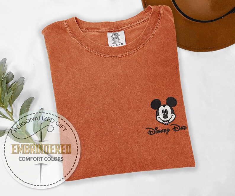 May include: Burnt orange Comfort Colors t-shirt with an embroidered Mickey Mouse design and the text "Disney Dad". The shirt has a classic crew neck and short sleeves. A brown hat is visible in the background.