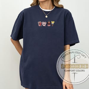 May include: A navy blue t-shirt with a white embroidered design of a popcorn bucket, a Mickey Mouse head, a donut, a Minnie Mouse head, and an ice cream cone. The shirt has a crew neck and short sleeves.