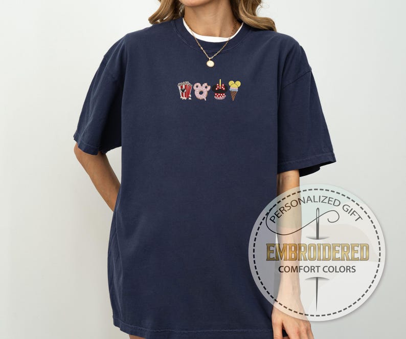 May include: A navy blue t-shirt with a white embroidered design of a popcorn bucket, a Mickey Mouse head, a donut, a Minnie Mouse head, and an ice cream cone. The shirt has a crew neck and short sleeves.