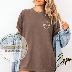 May include: A brown t-shirt with the text "Tired Moms Club" in white lettering. The shirt is being worn by a person with long brown hair. The person is wearing a denim jacket over the shirt.