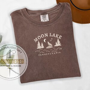 Pu&ograve; includere: Una maglietta marrone con un disegno ricamato bianco che dice "Moon Lake Pennsylvania" con un lupo che ulula alla luna e pini.