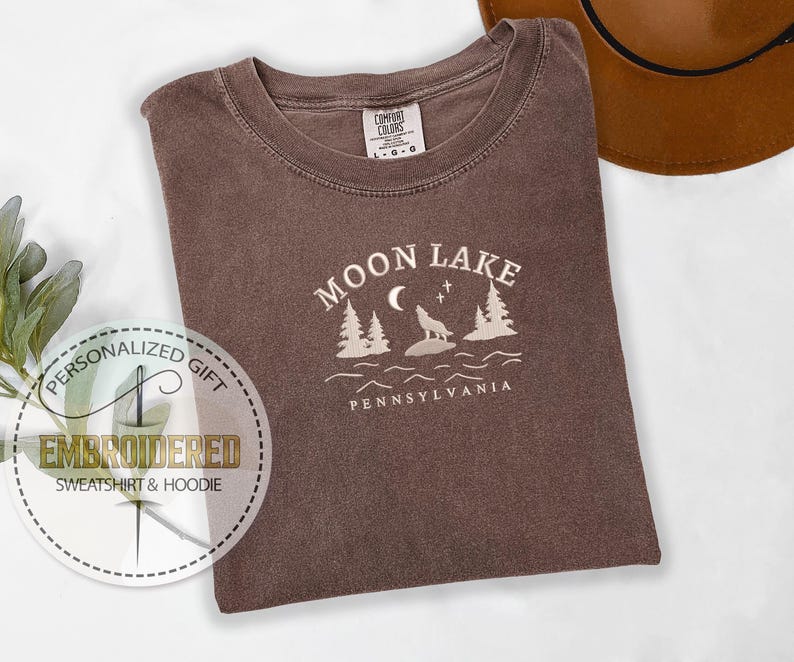 Pu&ograve; includere: Una maglietta marrone con un disegno ricamato bianco che dice "Moon Lake Pennsylvania" con un lupo che ulula alla luna e pini.