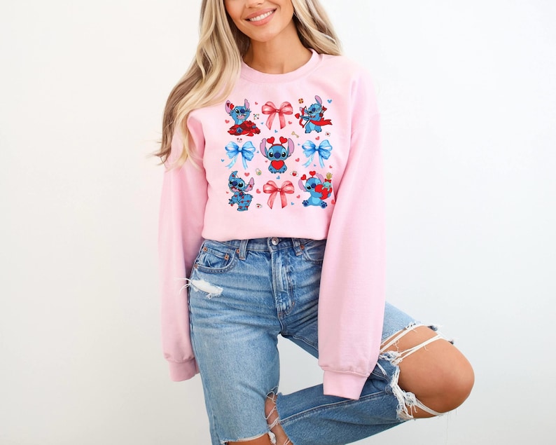 K&ouml;nnte beinhalten: Ein rosa Sweatshirt mit einem Grafikdruck von blauen und roten Comicfiguren mit Schleifen und Herzen.