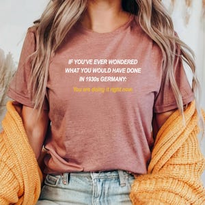 May include: A mauve t-shirt with the text "IF YOU'VE EVER WONDERED WHAT YOU WOULD HAVE DONE IN 1930s GERMANY: You are doing it right now." printed in white.