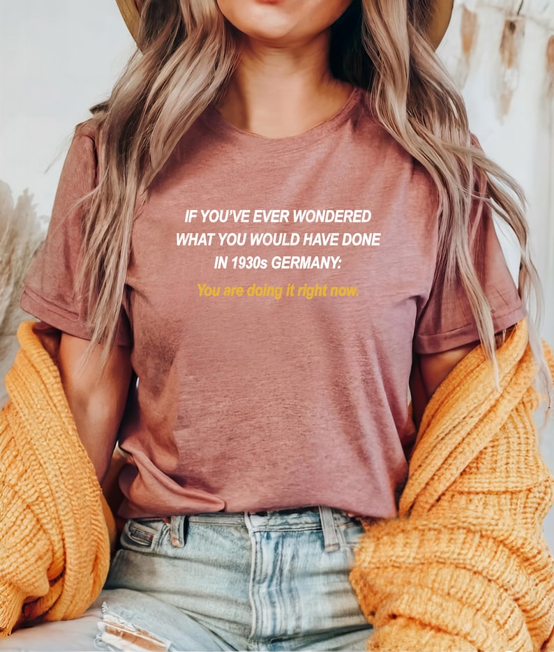 May include: A mauve t-shirt with the text "IF YOU'VE EVER WONDERED WHAT YOU WOULD HAVE DONE IN 1930s GERMANY: You are doing it right now." printed in white.