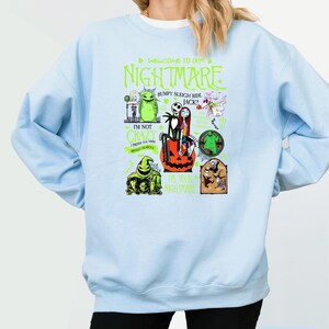May include: Pale blue sweatshirt with a graphic design featuring characters and text from "The Nightmare Before Christmas." The design includes the words "WELCOME TO OUR NIGHTMARE" and other phrases. The sweatshirt is a light blue colour.
