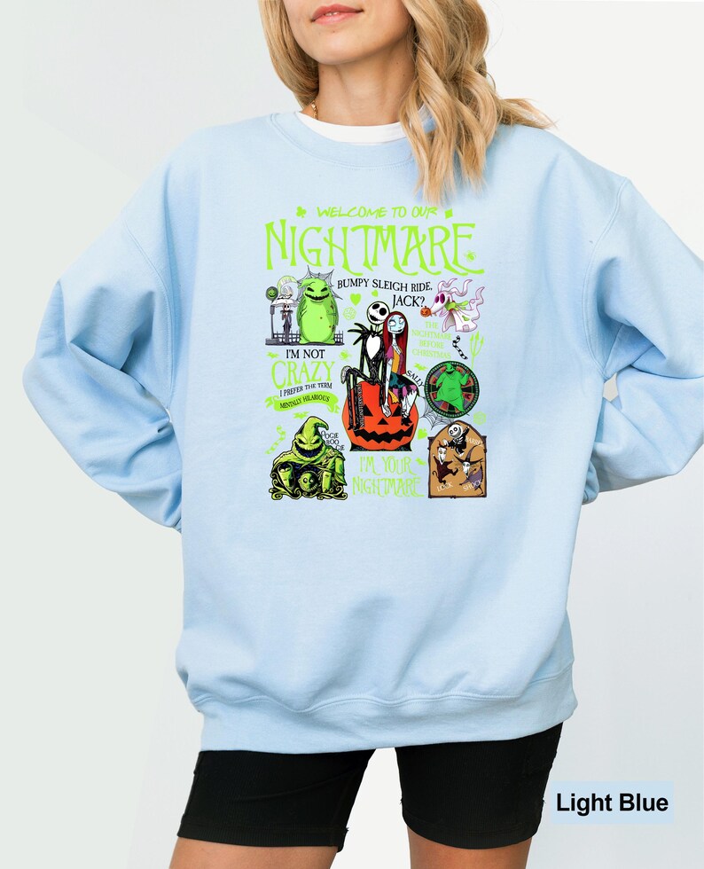 May include: Pale blue sweatshirt with a graphic design featuring characters and text from "The Nightmare Before Christmas." The design includes the words "WELCOME TO OUR NIGHTMARE" and other phrases. The sweatshirt is a light blue colour.