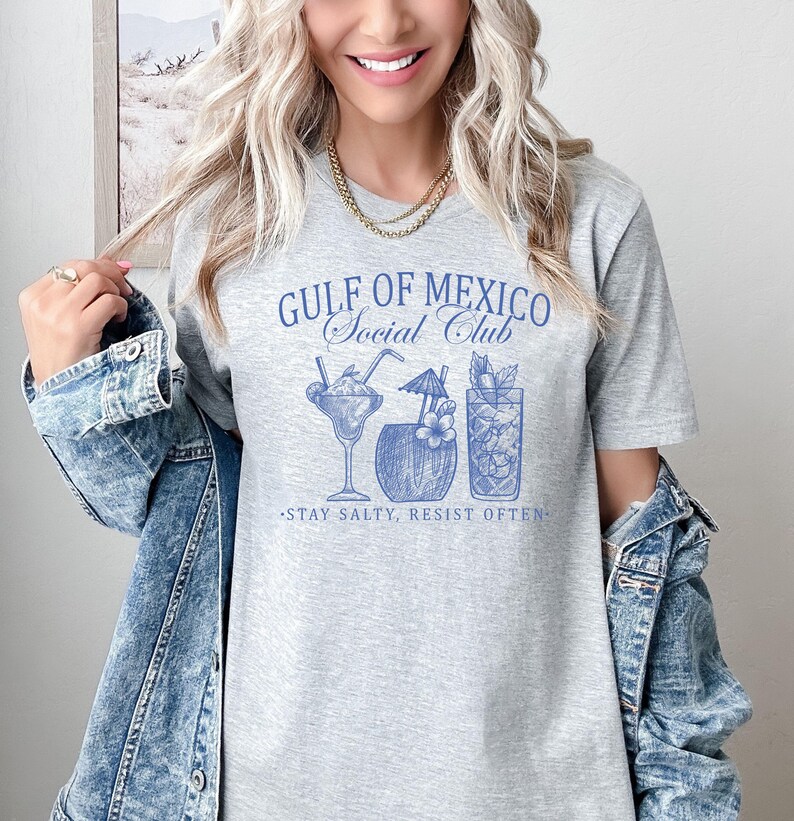 Puede incluir: Una camiseta gris con un dise&ntilde;o gr&aacute;fico azul que dice "Gulf of Mexico Social Club" y presenta tres bebidas de c&oacute;ctel. El texto debajo del gr&aacute;fico dice "Stay Salty, Resist Often."