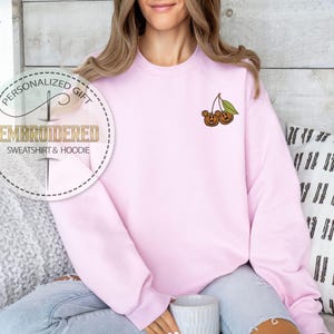 May include: A light pink sweatshirt with an embroidered design of two cherries with a green leaf. The sweatshirt has the text "EMBROIDERED SWEATSHIRT & HOODIE" in a circular design. The sweatshirt is a personalized gift.