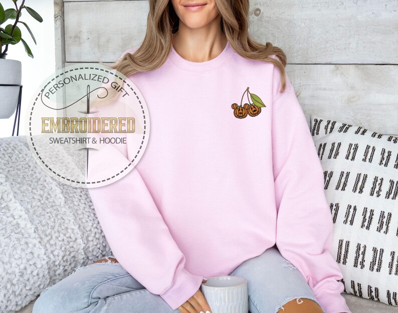 May include: A light pink sweatshirt with an embroidered design of two cherries with a green leaf. The sweatshirt has the text "EMBROIDERED SWEATSHIRT & HOODIE" in a circular design. The sweatshirt is a personalized gift.