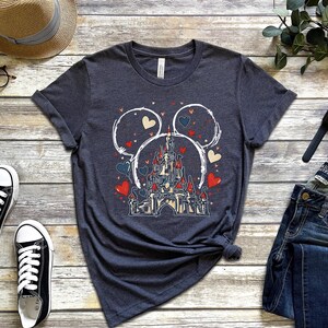 May include: Dark gray t-shirt featuring a castle design within Mickey Mouse ears, with red, blue, and beige hearts. The shirt is paired with black sneakers, a straw hat, and blue jeans. A watch and glasses are also visible.