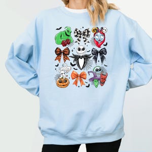 May include: Pale blue sweatshirt with a pattern of cartoon character bows, including Jack Skellington and Sally. The design is surrounded by bats and spiderwebs. The text "Light Blue" is at the bottom of the image.