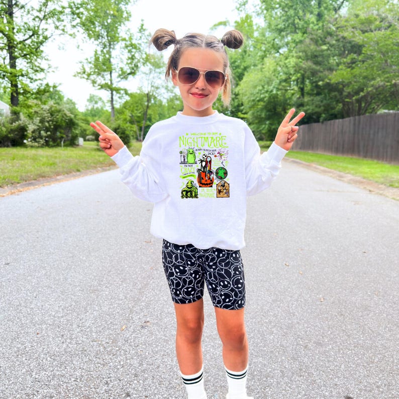 May include: A white sweatshirt featuring a graphic from "The Nightmare Before Christmas" and black biker shorts with a white smiley face pattern. The child is wearing sunglasses and white socks with black stripes.